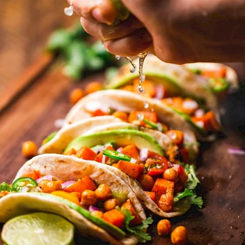 A hand squeezes lime over chickpea and vegetable tacos garnished with cilantro, on a wooden board with lime slices.