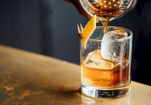 A classic cocktail being poured into a glass with ice and a citrus peel garnish on a wooden surface.