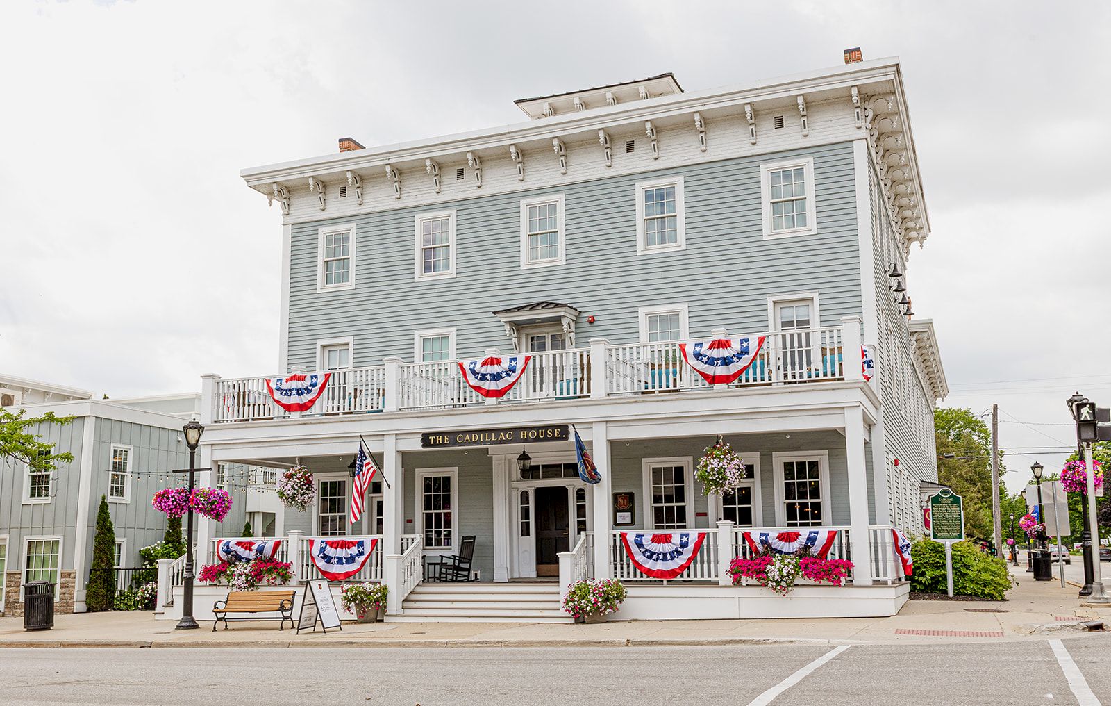 A white historic building with balcony bunting, flowers, and a sign reading &ldquo;The Cadillac House&rdquo; stands under a cloudy sky.