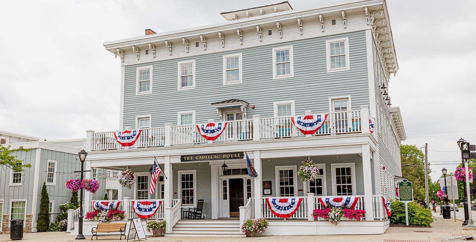 A white historic building with balcony bunting, flowers, and a sign reading &ldquo;The Cadillac House&rdquo; stands under a cloudy sky.