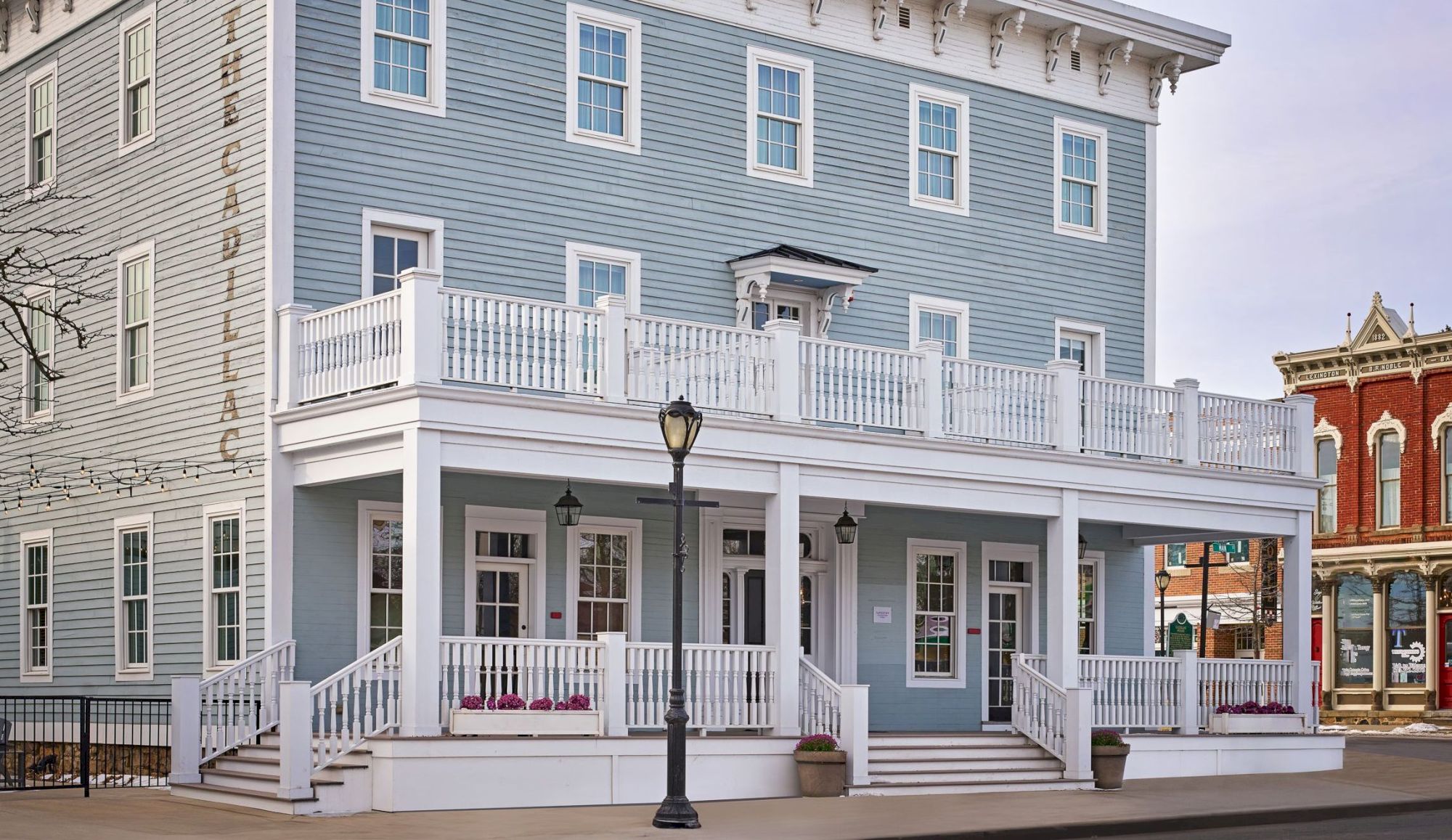 A large, light blue historic building with white trim and a porch, located on a corner in a quaint town setting.
