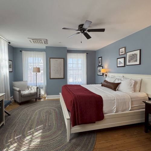 A bright bedroom with a bed, ceiling fan, bedside lamps, framed art, and sheer curtains by large windows.