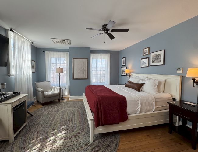 A bright bedroom with a bed, ceiling fan, bedside lamps, framed art, and sheer curtains by large windows.