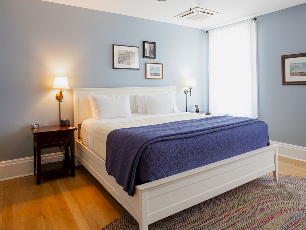 A neat bedroom with a white bed, bedside lamps, framed wall art, a ceiling fan, and a patterned rug on the floor.