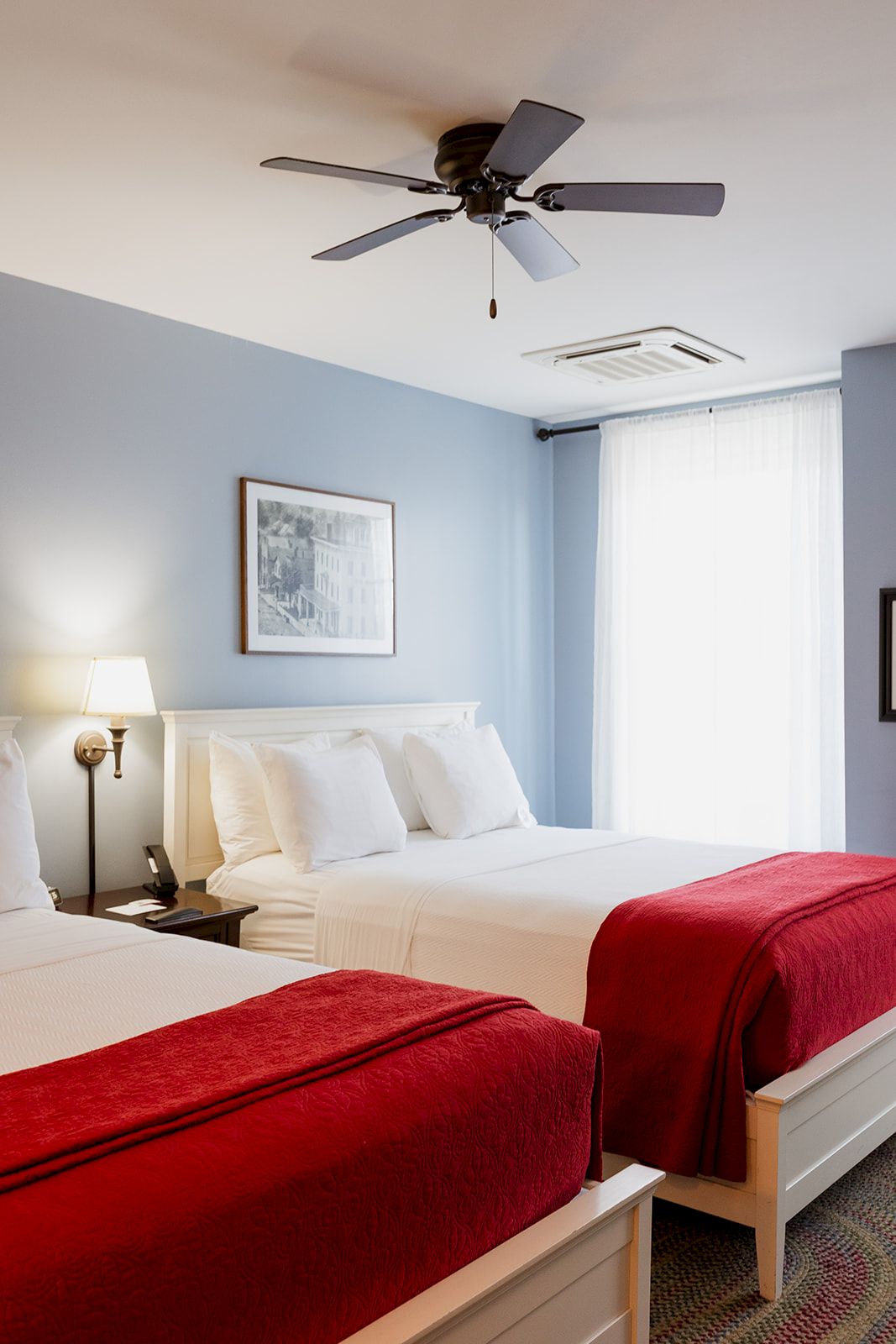 A bright hotel-style bedroom with two beds, red throws, and a ceiling fan above.