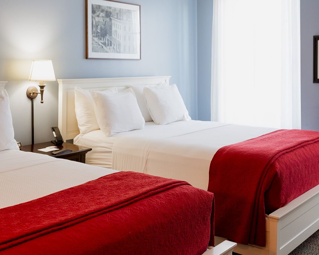 A bright hotel-style bedroom with two beds, red throws, and a ceiling fan above.
