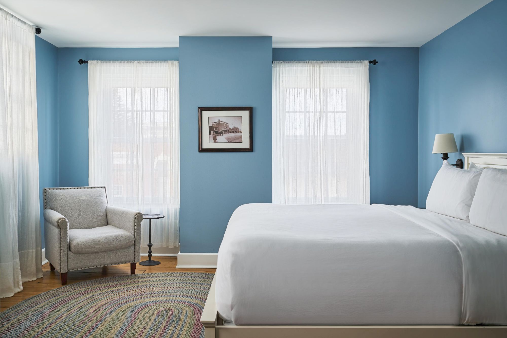 A serene bedroom with blue walls features a bed, armchair, small round table, wall art, and a woven rug, all bathed in natural light.