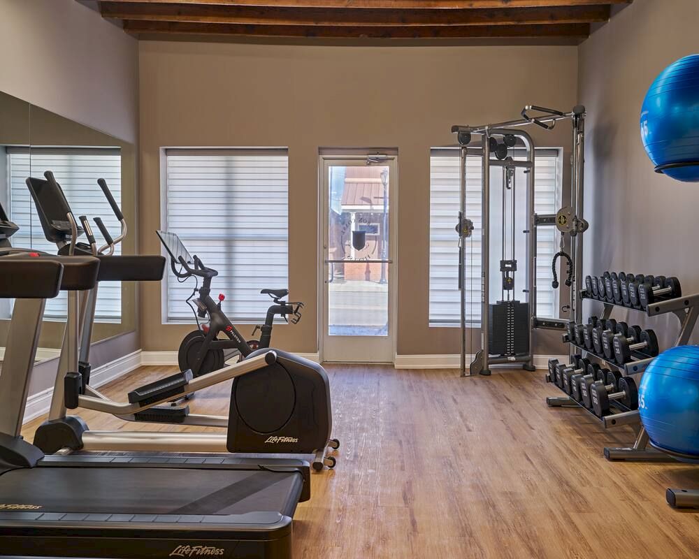 This image shows a gym with treadmills, exercise bikes, weights, a cable machine, and exercise balls. The room has a wooden floor.