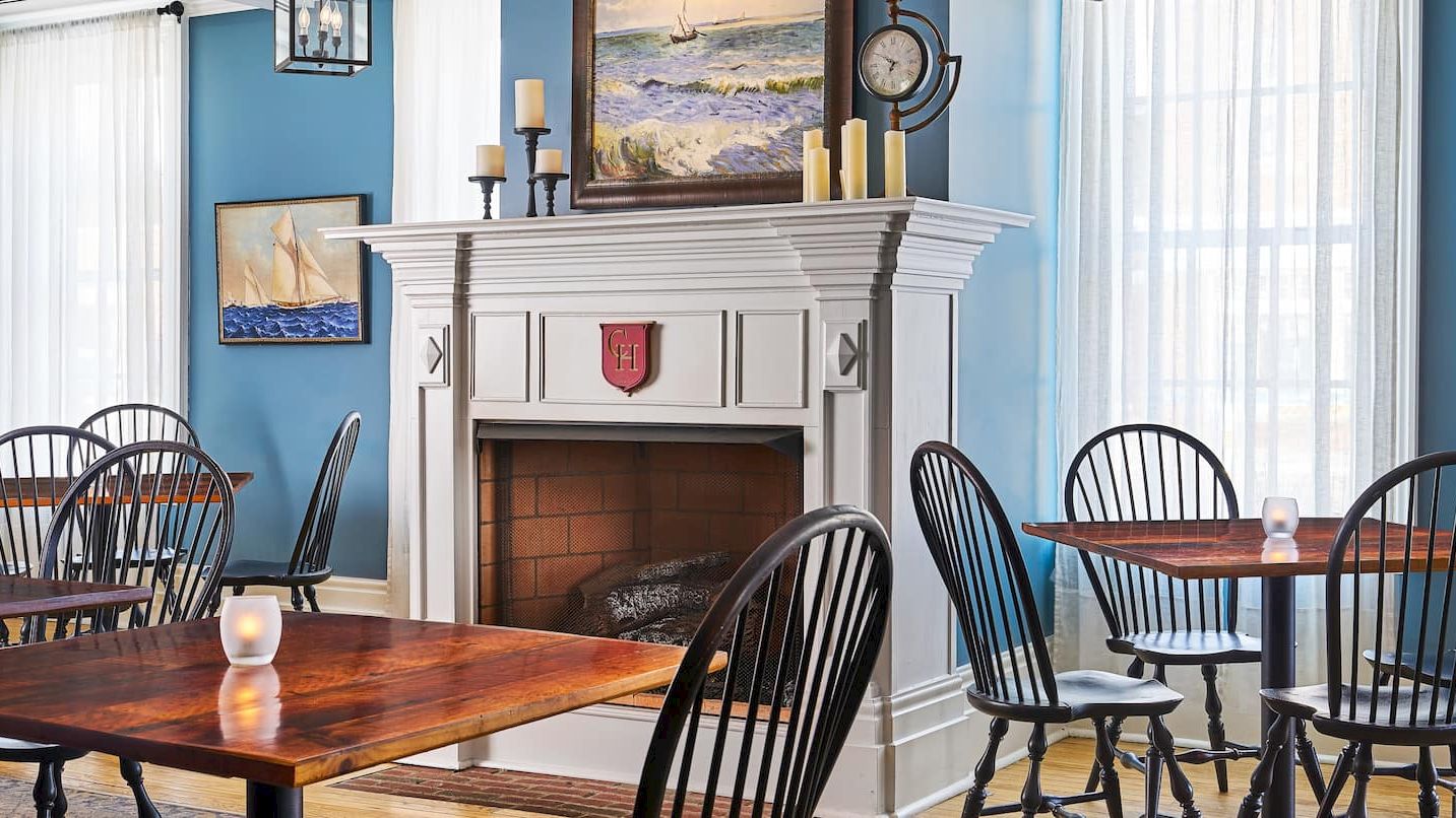 A cozy dining room with a fireplace, blue walls, wooden tables, and black chairs, featuring nautical-themed paintings on the walls.
