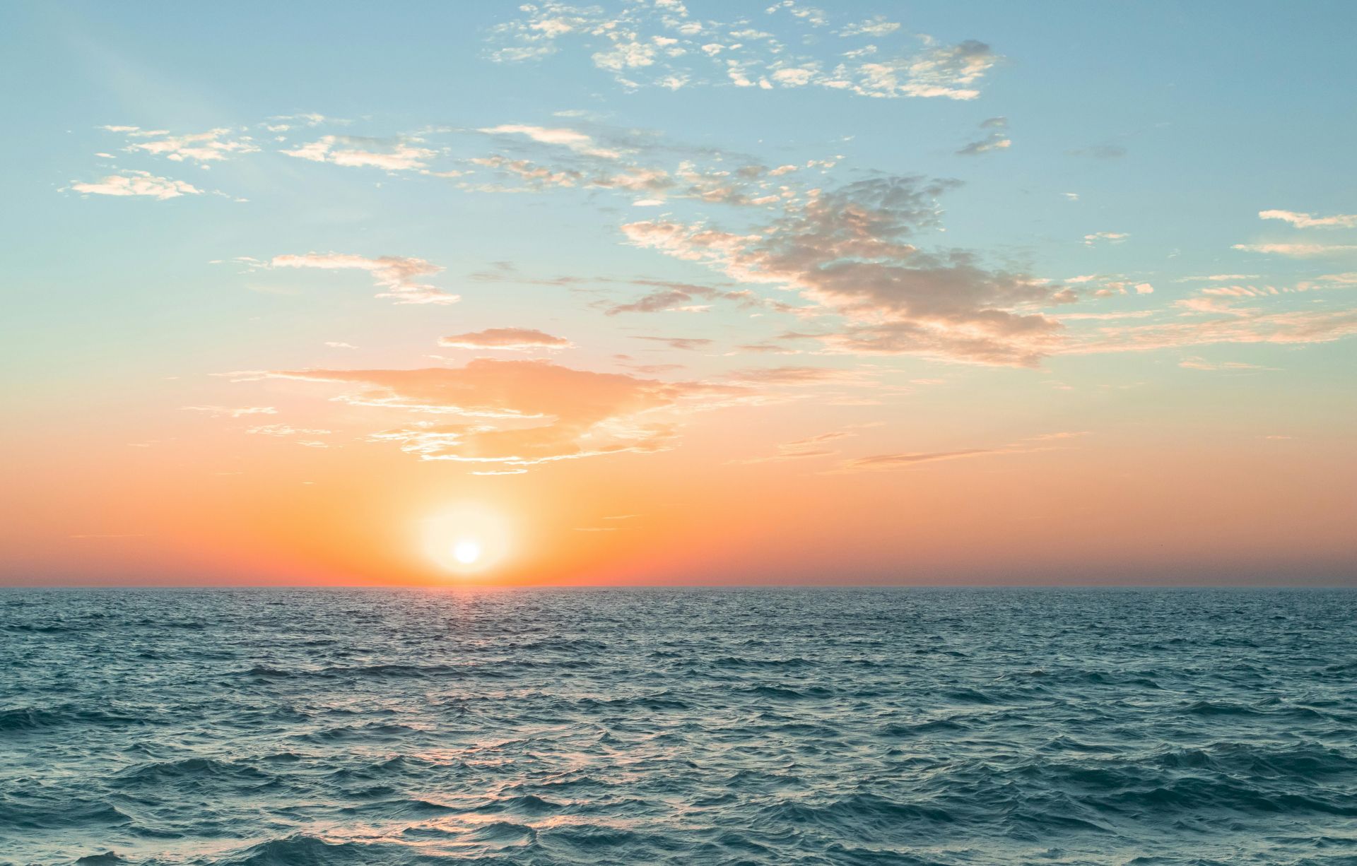 A serene ocean sunset with a bright sun near the horizon, fluffy clouds, and a tranquil sea under a colorful sky.