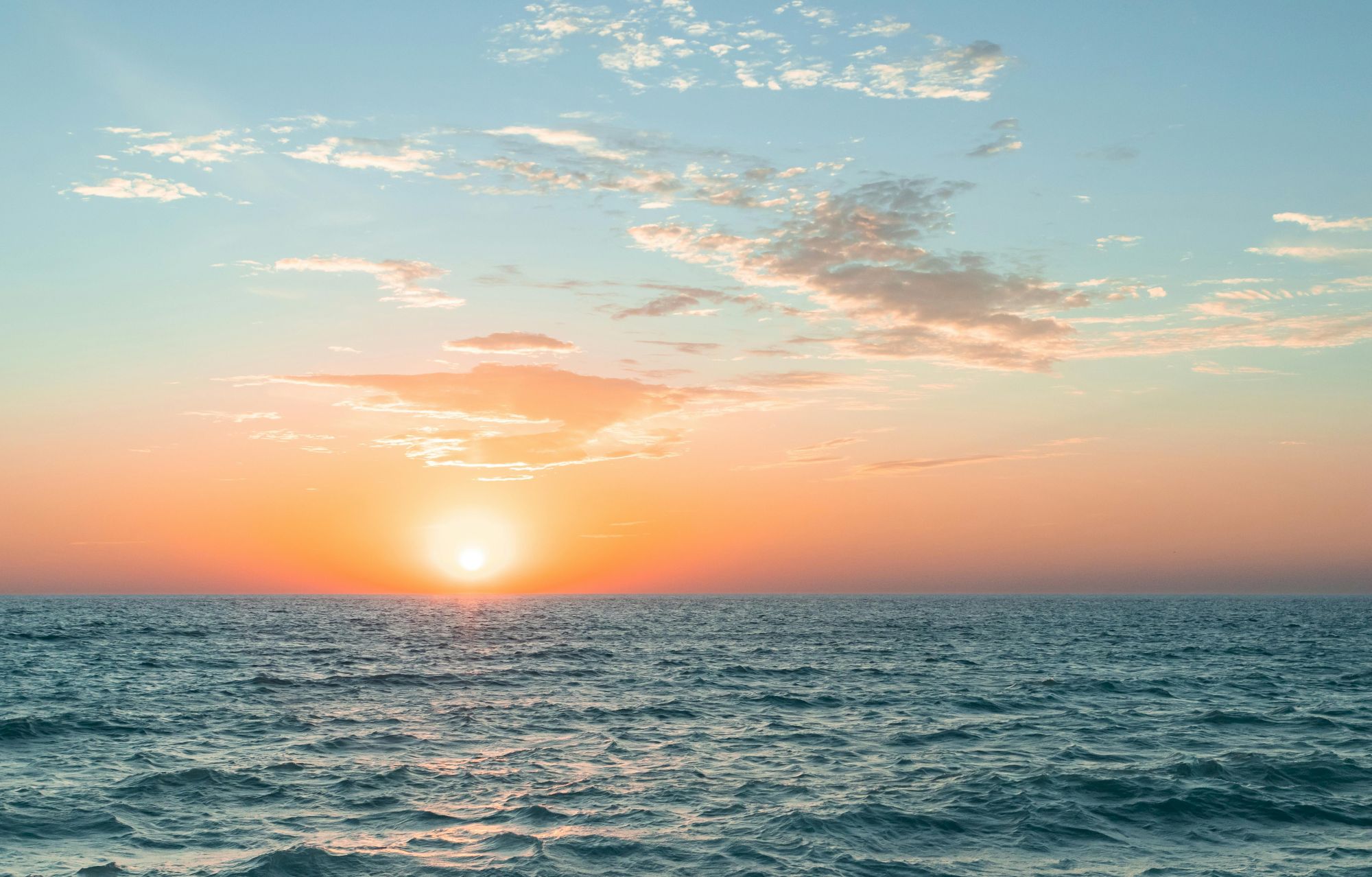 A serene ocean sunset with a bright sun near the horizon, fluffy clouds, and a tranquil sea under a colorful sky.