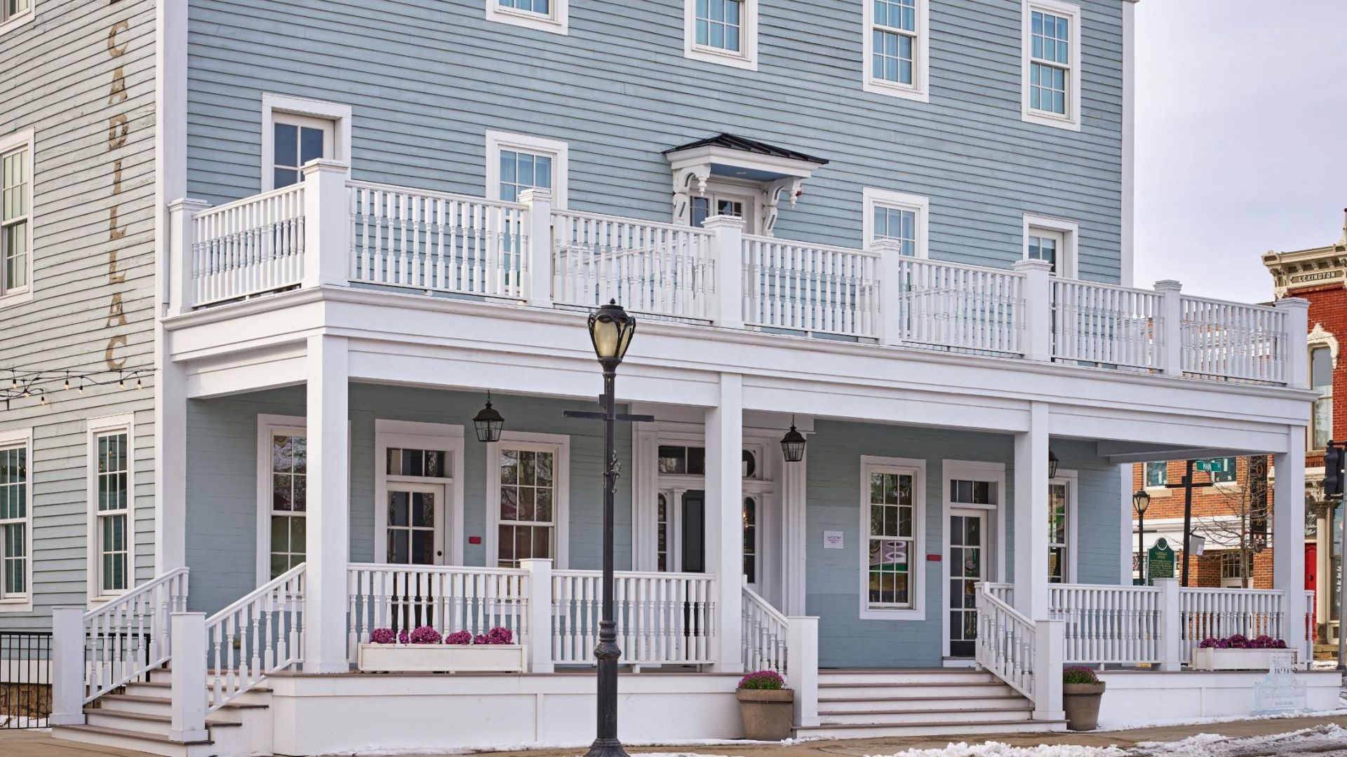A light blue building with a wraparound porch, white railings, and a lamp post stands beside a street in a small town.