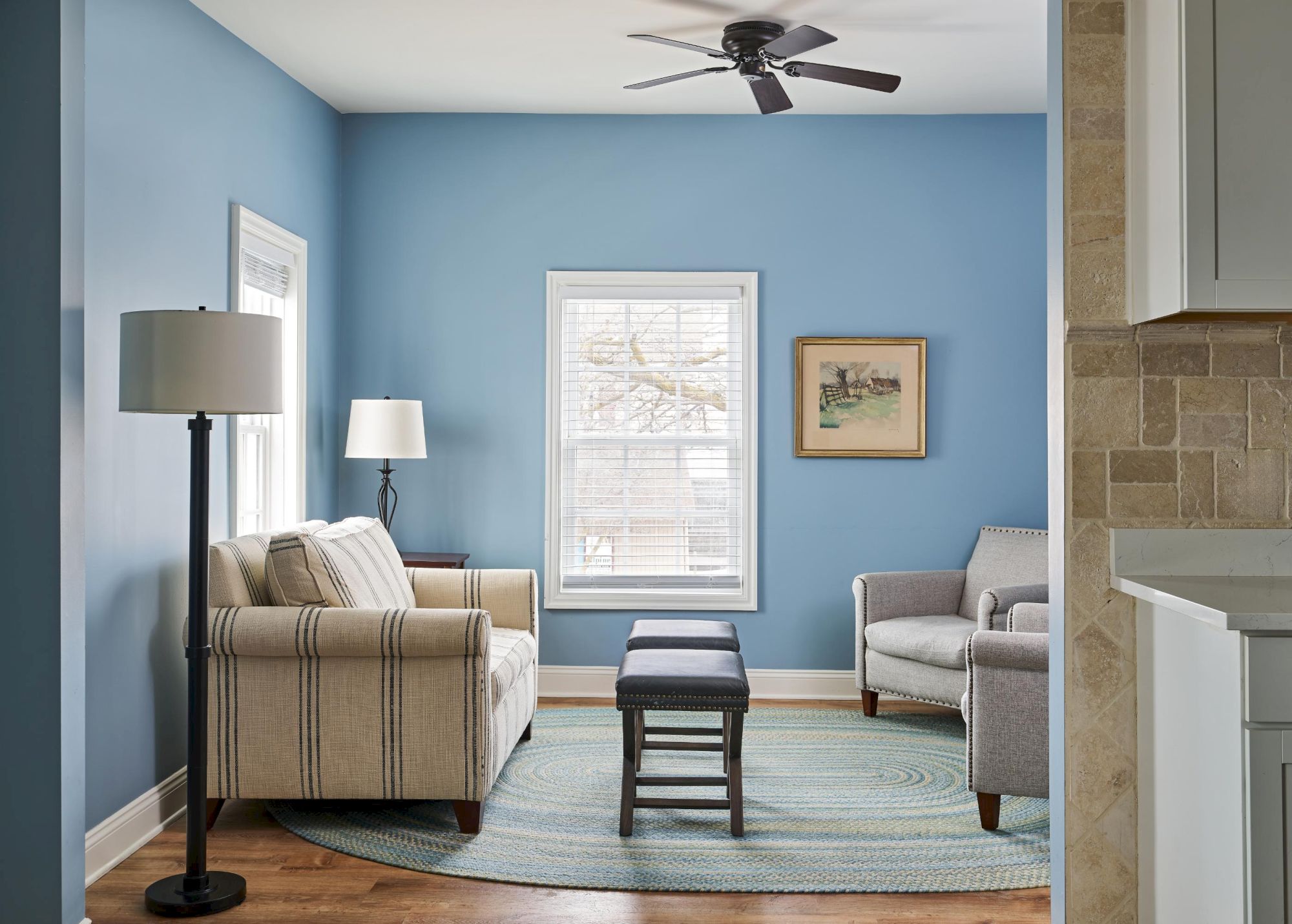 A cozy living room with blue walls, a striped sofa, armchair, floor lamp, and a rug. It has a ceiling fan and a window with blinds.
