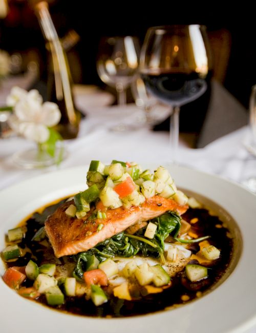 A dish featuring salmon topped with a cucumber-tomato mix, served on greens with a dark sauce, alongside wine glasses in a restaurant setting.