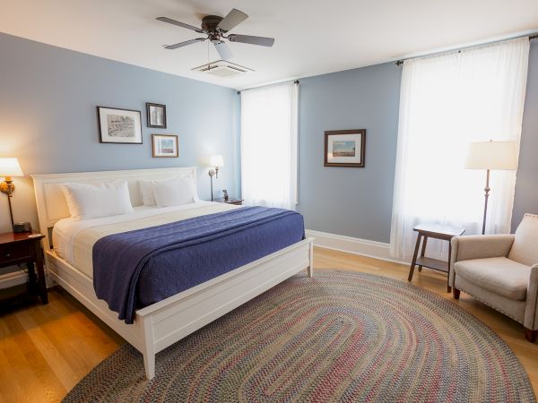 A bright blue-walled bedroom with a white bed, lamps, ceiling fan, framed art, and sheer curtains.