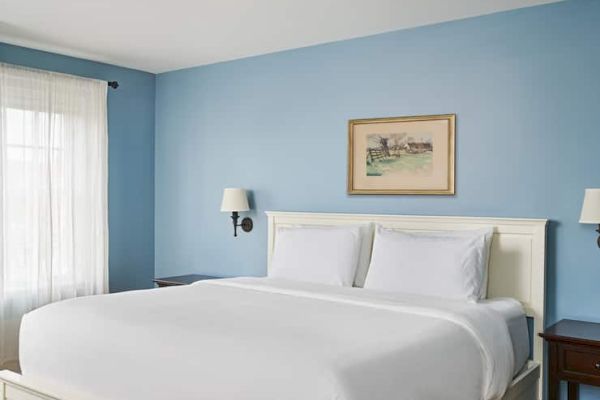 A bedroom with a white bed, blue walls, framed artwork, two wall lamps, and sheer curtains letting in natural light.