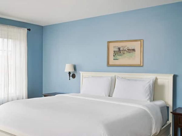 A bedroom with a white bed, blue walls, framed artwork, two wall lamps, and sheer curtains letting in natural light.