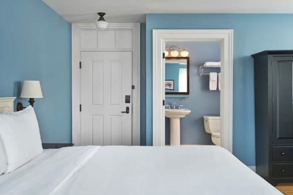 A cozy bedroom with blue walls, a white bed, and an open bathroom door showing a sink, mirror, and toilet.