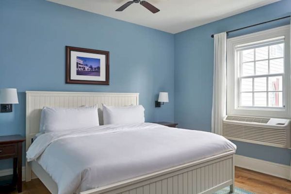 A cozy bedroom with a white bed, blue walls, wall sconces, a framed picture, ceiling fan, window with curtain, and air conditioning unit.