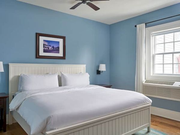 A cozy bedroom with a white bed, blue walls, wall sconces, a framed picture, ceiling fan, window with curtain, and air conditioning unit.