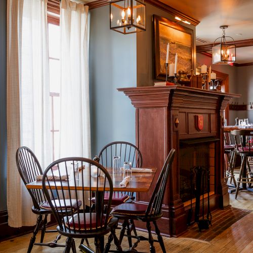 A cozy dining room with wooden chairs, a table set with glasses, and a fireplace mantel lit by hanging lanterns.