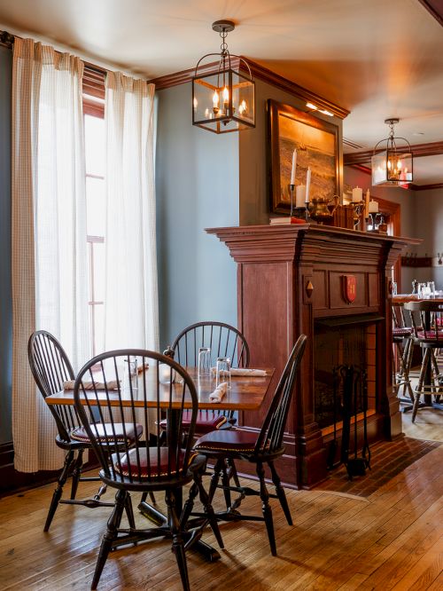 A cozy dining room with wooden chairs, a table set with glasses, and a fireplace mantel lit by hanging lanterns.
