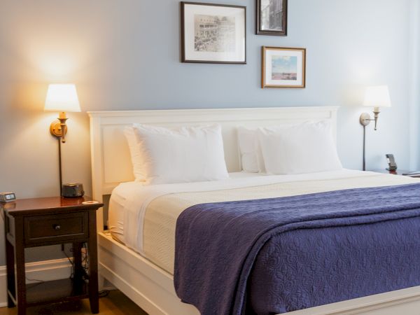 A tidy bedroom with a white bed, purple blanket, bedside lamps, and framed wall art under a ceiling fan.