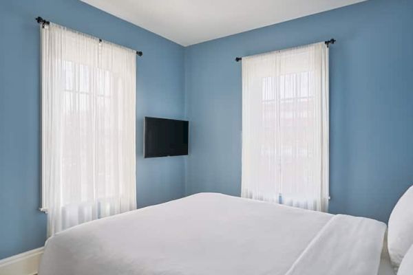 A bedroom with light blue walls, a bed, sheer curtains on two windows, and a wall-mounted TV.