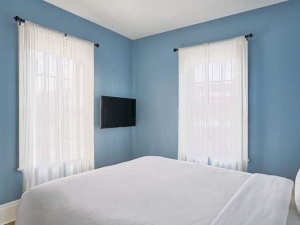 A bedroom with light blue walls, a bed, sheer curtains on two windows, and a wall-mounted TV.