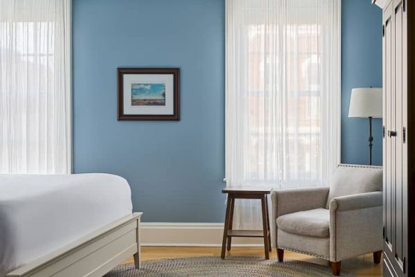 A bedroom with blue walls, white bed, armchair, small table, floor lamp, framed art, and light streaming through sheer curtains.