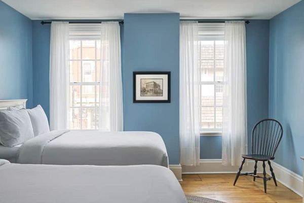 A blue-walled bedroom with two beds, white curtains, a framed picture, and a wooden chair by the window completes the scene.