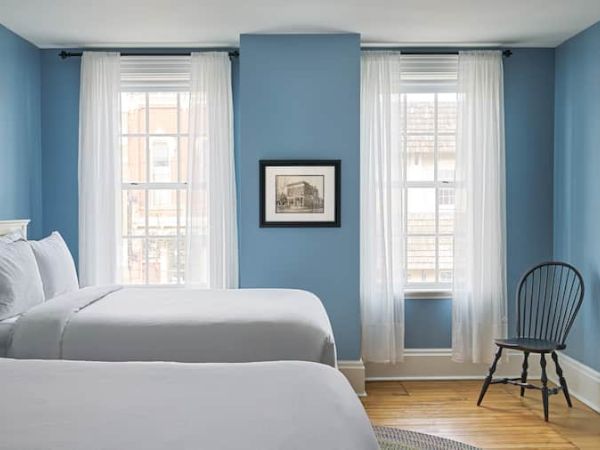 A blue-walled bedroom with two beds, white curtains, a framed picture, and a wooden chair by the window completes the scene.