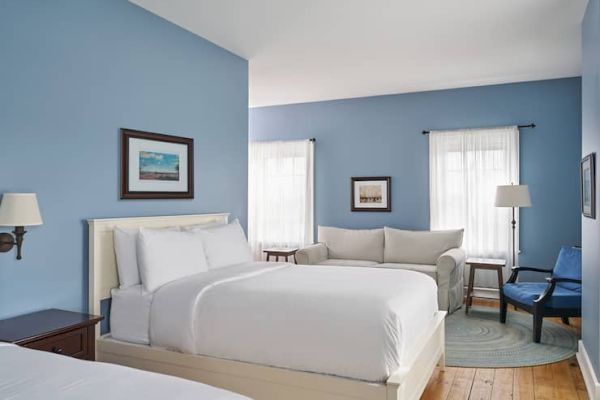 The image shows a bedroom with blue walls, white bedding, a sofa, two windows with curtains, a lamp, and framed art on the walls.