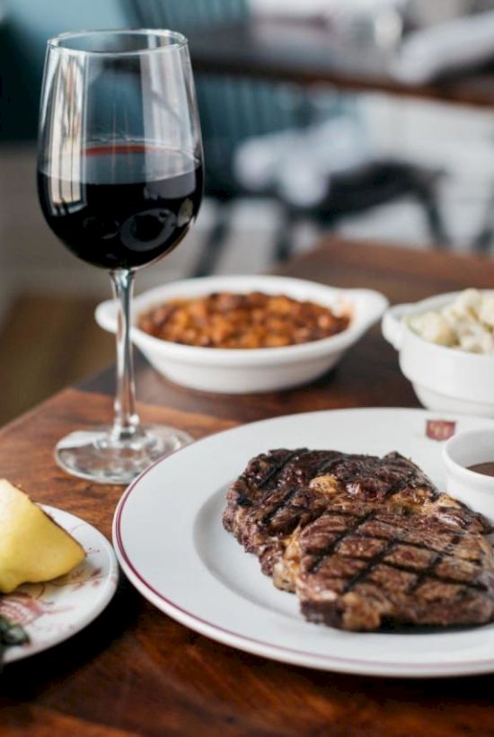 A steak with sauce, asparagus with lemon, baked beans, creamy salad, and a glass of red wine on a wooden table.