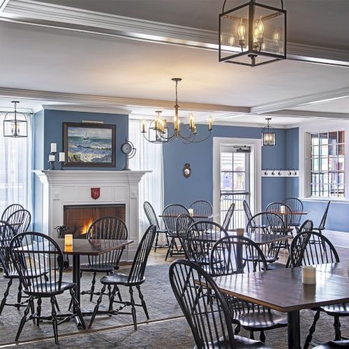 The image shows a dining area with wooden tables and chairs, a blue wall, a lit fireplace, chandeliers, and framed artwork on the walls.
