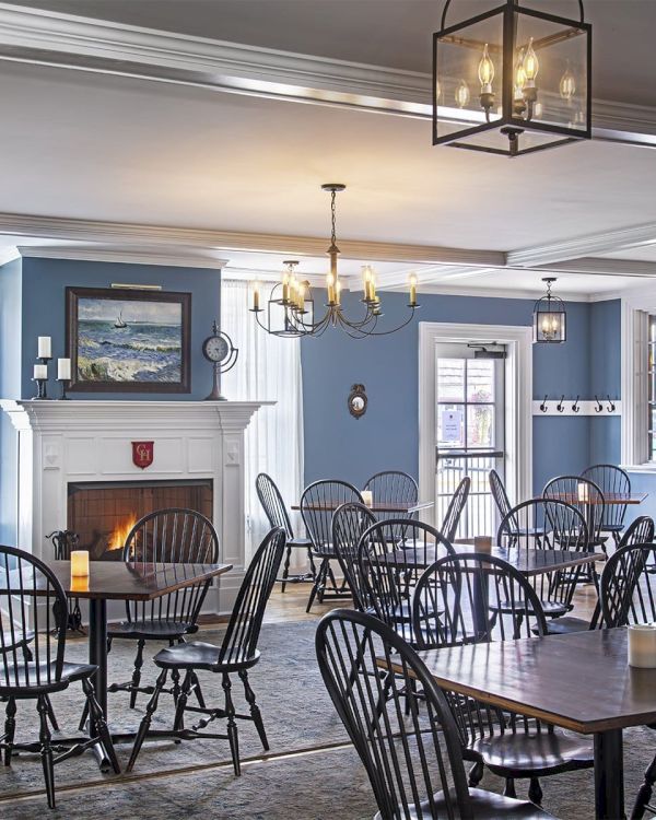 The image shows a dining area with wooden tables and chairs, a blue wall, a lit fireplace, chandeliers, and framed artwork on the walls.