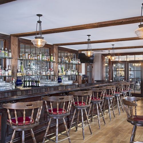 A stylish bar with wooden stools, a variety of bottles on shelves, and hanging pendant lights, creating a warm, inviting atmosphere.