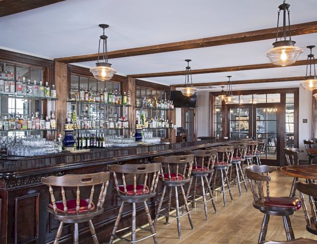 A stylish bar with wooden stools, a variety of bottles on shelves, and hanging pendant lights, creating a warm, inviting atmosphere.