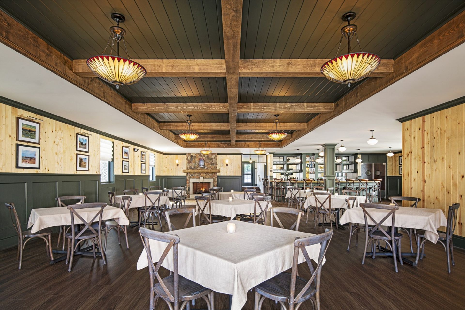 A warmly lit, spacious restaurant with wooden beams, chandeliers, and neatly arranged tables and chairs on a wooden floor.