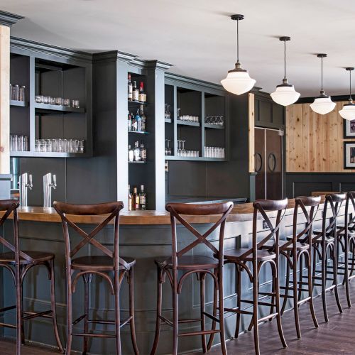 This image shows a bar with wooden stools, a counter, shelves with glasses and bottles, and hanging lights.