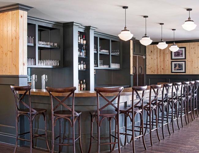 This image shows a bar with wooden stools, a counter, shelves with glasses and bottles, and hanging lights.