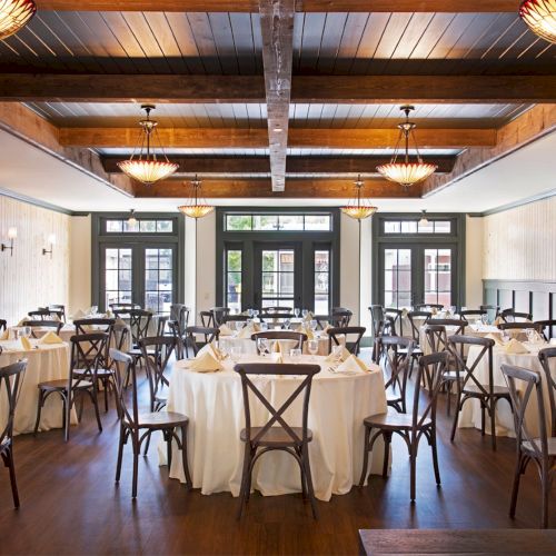 A spacious restaurant with round tables covered in white cloths, surrounded by wooden chairs under a beamed ceiling with elegant lighting.