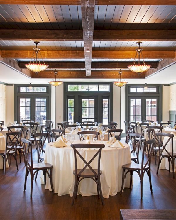 A spacious restaurant with round tables covered in white cloths, surrounded by wooden chairs under a beamed ceiling with elegant lighting.