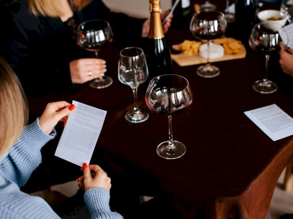 People sit around a table with wine glasses, a bottle, papers, and a cheese board, suggesting a formal or social gathering.
