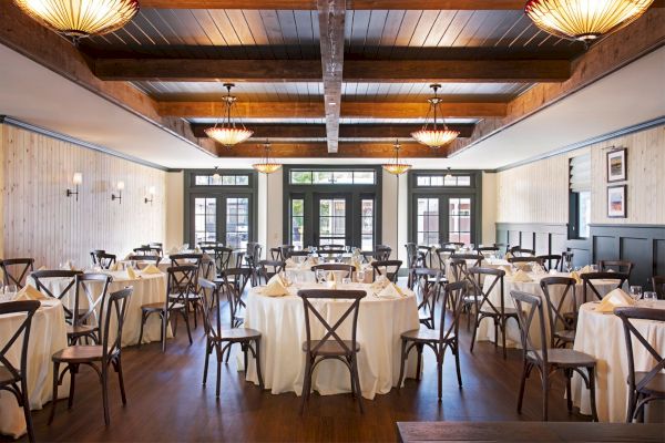 An elegantly set dining room features round tables with white tablecloths, wooden chairs, and warm lighting from chandeliers above.