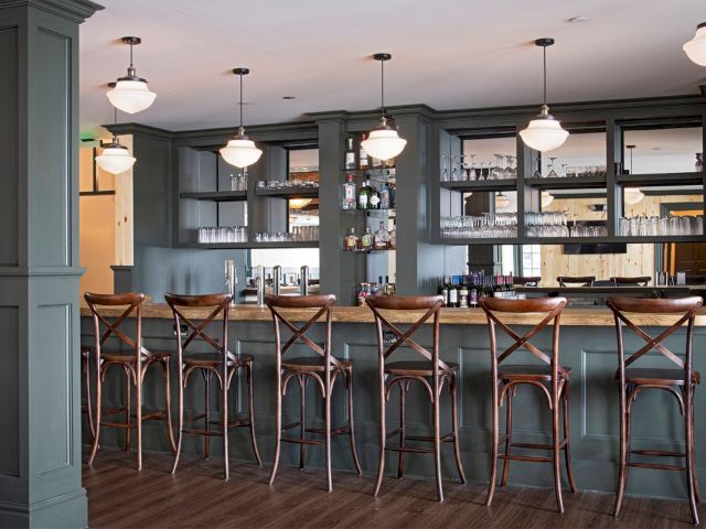 A stylish bar with six wooden chairs, hanging lights, a well-stocked shelf with glasses and bottles, and a wooden countertop.
