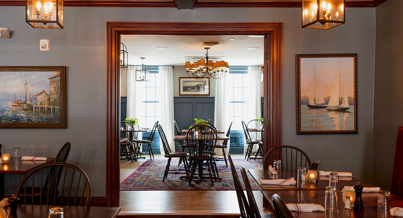 A warm dining room with wooden chairs, candlelit lanterns, framed nautical art, and a chandelier over the table.