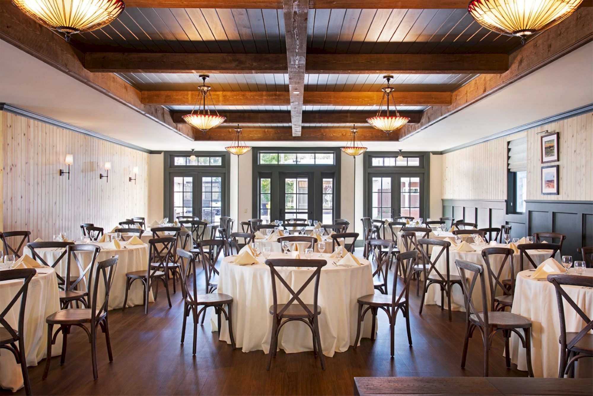 The image shows an elegant restaurant dining room with round tables, white tablecloths, wooden chairs, and decorative lights.