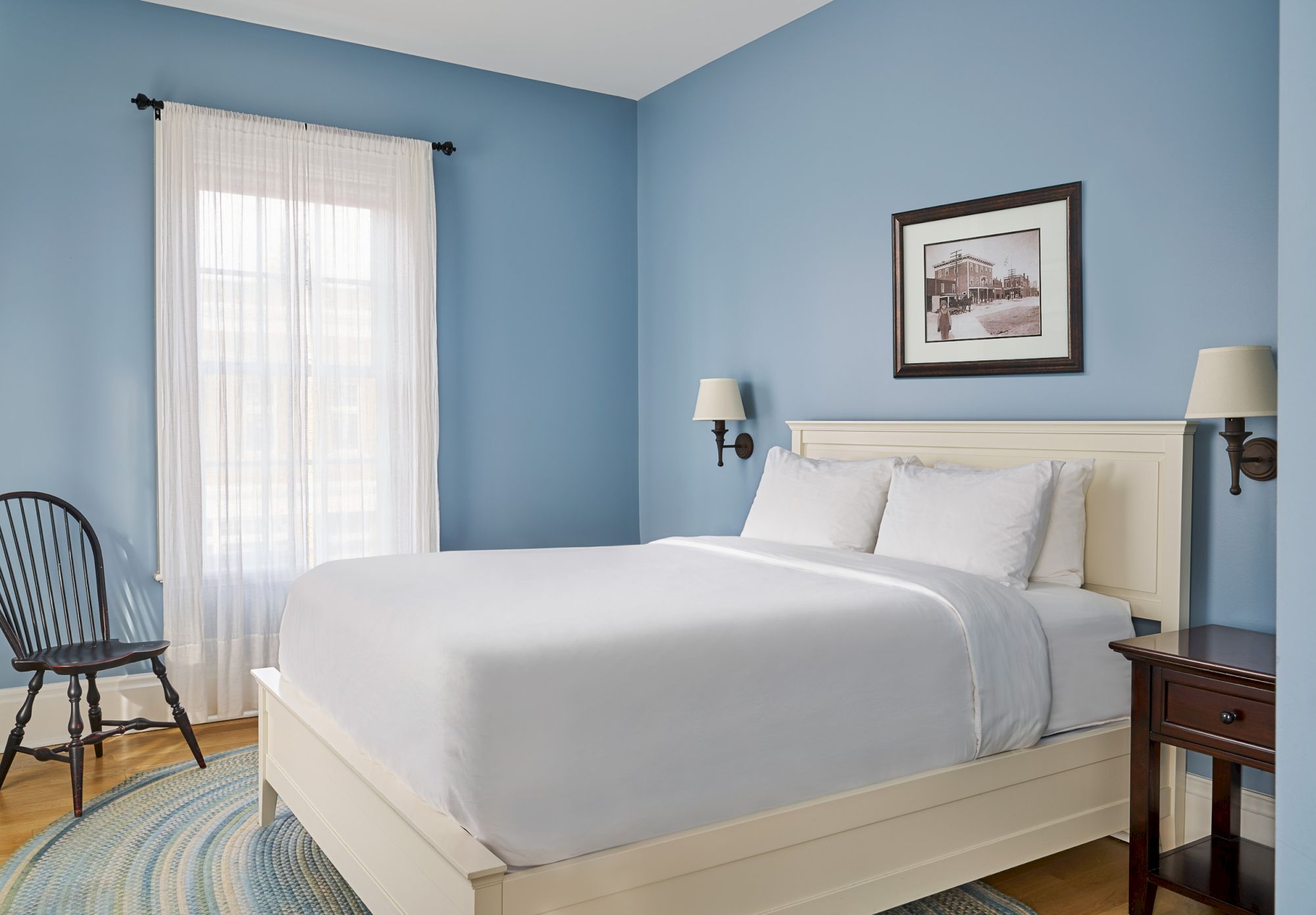 A neatly made bed in a blue-walled room with a framed photo, wall sconces, and a wooden chair by a window with sheer curtains.