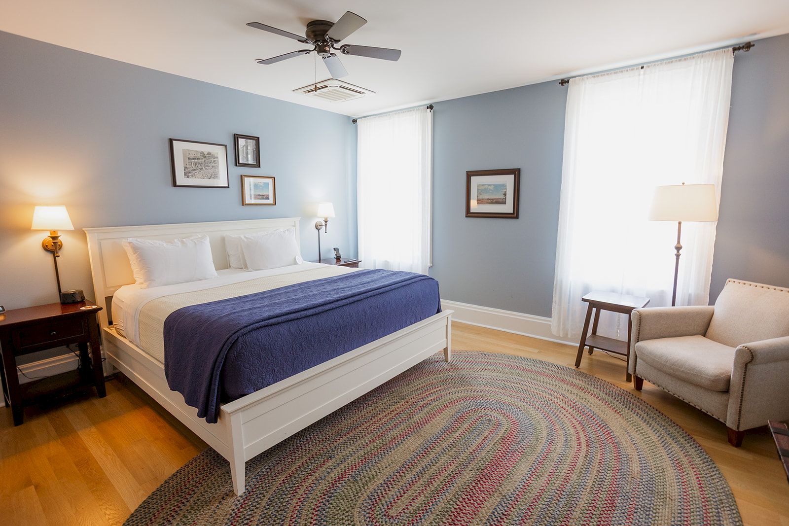 A bright bedroom with a white bed, blue bedding, ceiling fan, framed art, lamps, and sheer curtains over the windows.